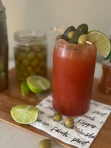 A photo of a bloody maria cocktail in the foreground on a napkin with a shaker cup, olives, limes and tajin in the background