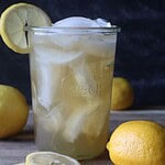 Photo of a glass of green tea lemonade with a lemon slice on the rim and slices of lemons and whole lemons on a cutting board in front of a black background
