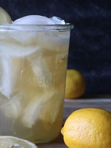 Photo of a glass of green tea lemonade with a lemon slice on the rim and slices of lemons and whole lemons on a cutting board in front of a black background