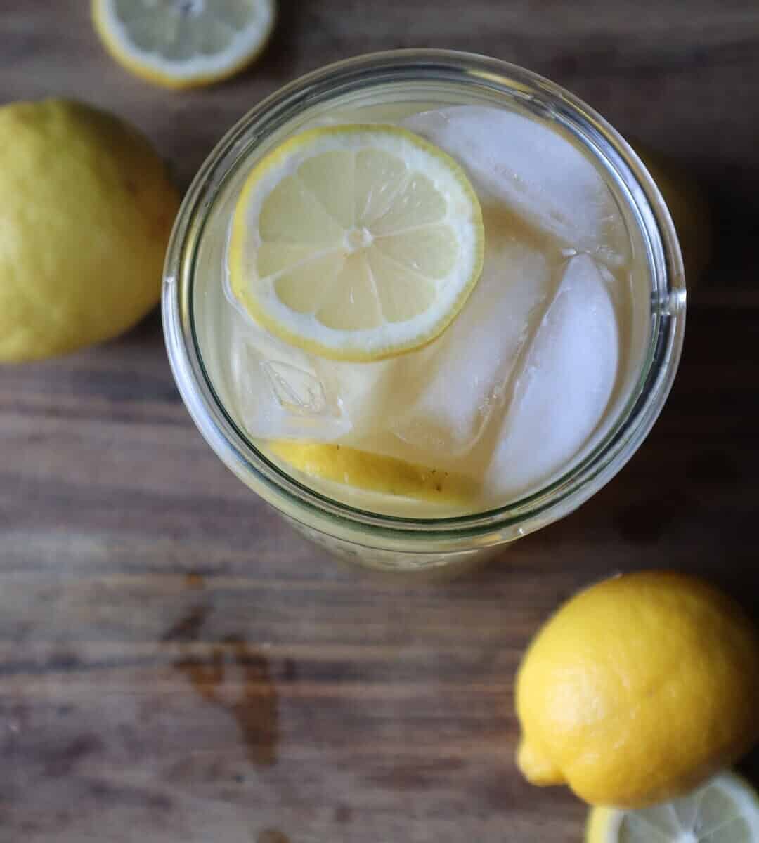 An aerial photo of a glass of green tea lemonade with a lemon slice on the rim and sliced lemons and whole lemons, all on a wooden cutting board in front of a black background