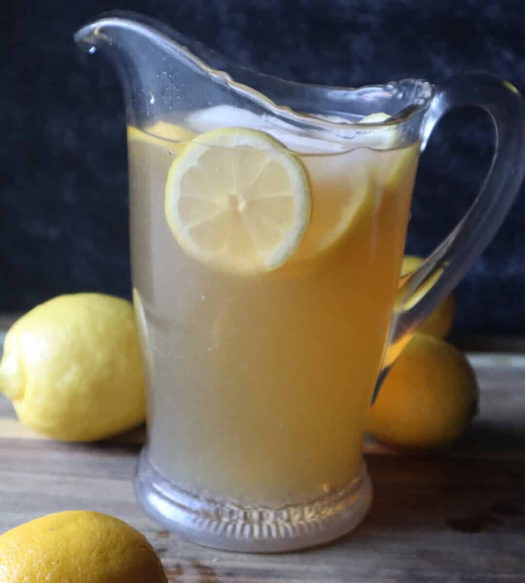 A photo of a pitcher of green tea lemonade and sliced lemons and whole lemons, all on a wooden cutting board in front of a black background