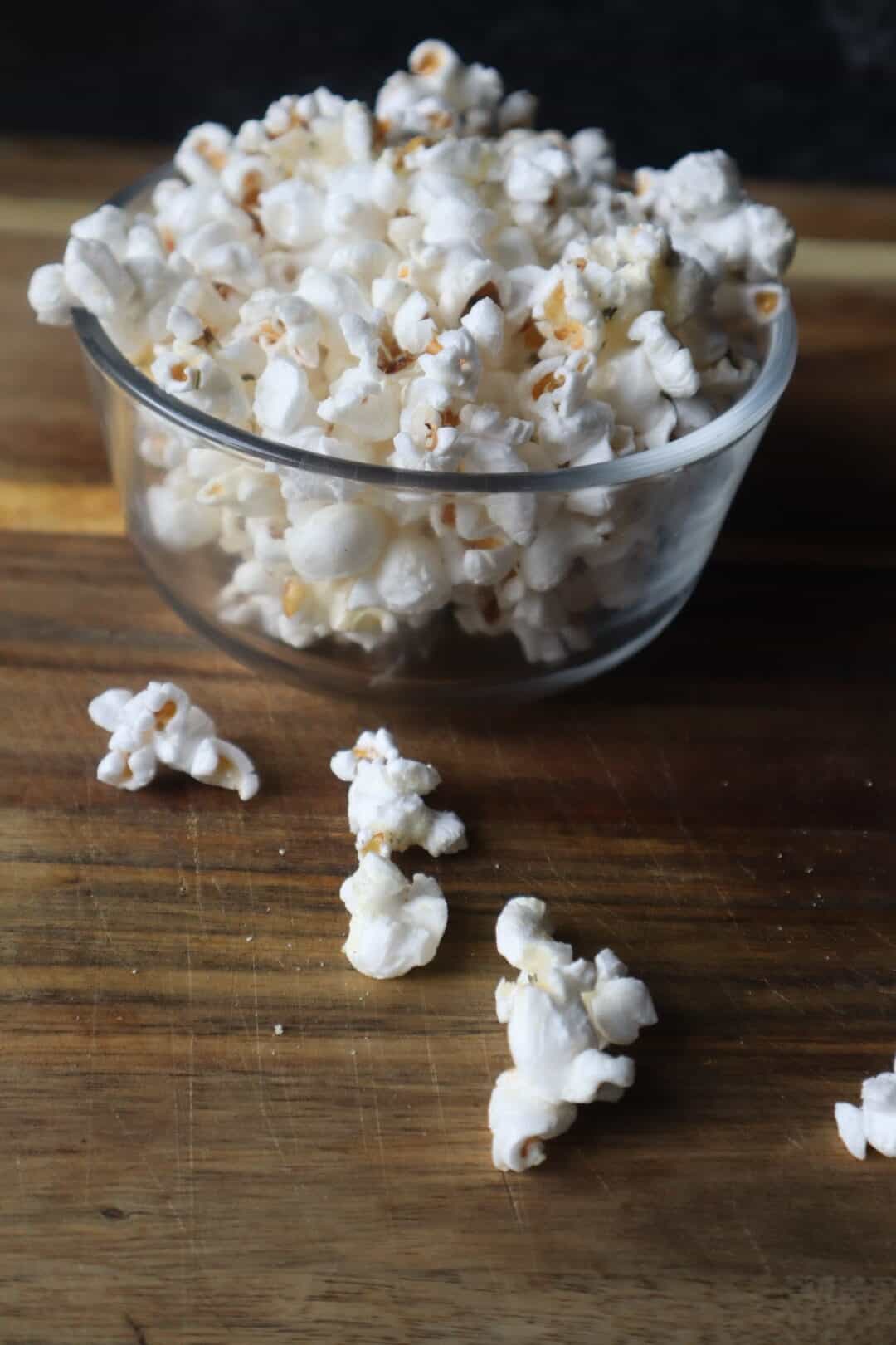 A clear glass bowl of popped popcorn with some kernels falling out onto the cutting board the bowl is resting on.