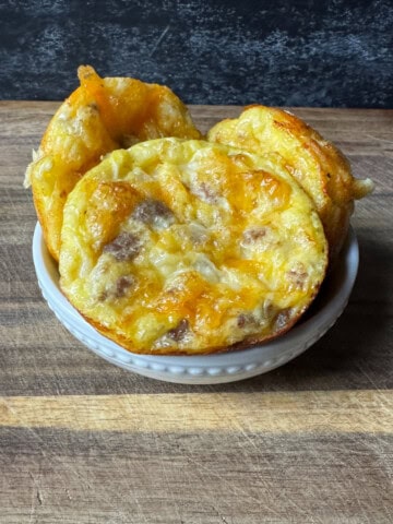 three sausage and cheese high protein egg bites in a white bowl sitting on a wooden cutting board in front of a black background